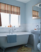 Naturfarben gestreifte Jalousie neben pastellblauer Wand und Klauenfüsse an freistehender Badewanne