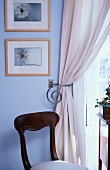 Antiker Stuhl vor Bildern auf hellblauer Wand und Vorhang mit Raffhalter aus Metall