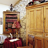 Kleine Sitzecke mit Tischdecke, Lampenbezug und ledergebundenen Bücher in Bordeauxrot neben antikem Holzschrank vor Natursteinmauer