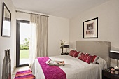 Pinkfarbener Überwurf und Kissen auf dem Bett mit grau gepolstertem Kopfende in einem Schlafzimmer mit weissen Vorhängen