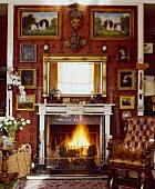 Bilder und ein goldgerahmter Spiegel über dem Kamin in einem roten Wohnzimmer mit antikem Lederrollstuhl