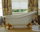 Eine frei stehende Badewanne mit Krallenfüssen vor dem Fenster mit weißem Rollo und Toile-de-Jouy Vorhängen