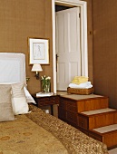 Gemusterte goldene Leinentücher auf dem Bett im hellbraunen Schlafzimmer mit Schubladen in den Stufen zur weißen Tür