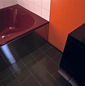 Eine dunkelrote Badewanne, schwarze Bodenfliesen und eine orange Holzplatte dominieren das moderne Badezimmer