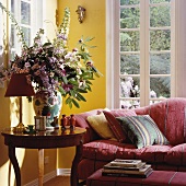Ecke in traditionellem Wohnzimmer mit violettem Blumenstrauss und rotem Polstersofa vor gelber Wand mit weissen Sprossenfenstern