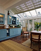 Wintergartenartige Vergrößerung einer Wohnküche mit pastellblauen Einbauschränken, Sisal-Matten auf Holzböden und antiken Holzsesseln mit Geflecht