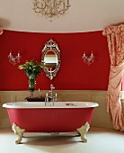 Ovaler, venezianischer Spiegel und Kristallleuchter an roter Wand hinter freistehender Badewanne, himbeerfarben mit weissen Klauenfüssen