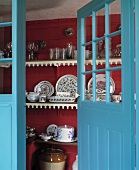 Blick durch eine blaue Holztür mit Sprossenfenstern in Regalraum mit gemustertem Geschirr und Gläsern