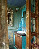 Blick durch historische Holzwand mit Butzenscheiben auf Waschbecken in antikem, blau gestrichenem Spiegelschrank