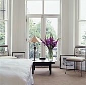 Eine Tischlampe und lila Gladiolen auf einem niedrigen Tisch vor dem französischen Fenster in einem weissen zeitgenössischen Schlafzimmer