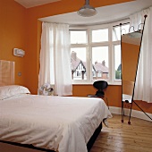 weiße Bettdecke auf dem Bett und weiße Vorhänge vor dem Erkerfenster in einem modernen orangefarbenen Schlafzimmer mit einem hohen freistehenden Spiegel