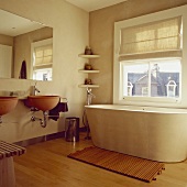 Ovale Betonbadewanne, Philippe Starck Waschbecken und Holzmatte auf dem Ahornboden in einem Badezimmer mit cremefarbener Fensterrollo