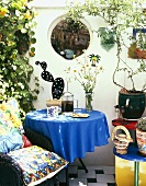 Bunter Terrassenplatz mit Kissen auf Stuhl im Ethnostil und Tisch mit blauem Tischtuch vor weisser Wand mit rundem Fensterausschnitt