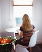 A blonde woman working at a laptop with a bowl of tomatoes in the foreground