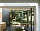 Dining table and side board of light wood in front of a bank of windows with a garden view