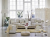 Modernes Wohnzimmer mit zimmerhoher, großer Fensterfront, weißem Megasofa, Glastisch und einem filigranen, weissen Paravent im arabischen Stil