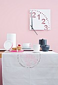 A table laid with tea crockery and cakes against a pink wall with a modern clock