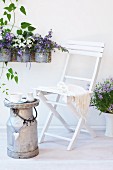 A white wooden chair and a vintage milk churn with flowerpots of campanula and petunias hanging on the wall