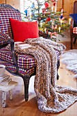A knitted blanket, a cushion and a book on an armchair with a Christmas tree in the background