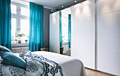 A white wardrobe with wide sliding elements and a mirrored front in a bedroom with blue curtains
