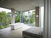 Corner of a sleeping area with free standing bathtub in front of a panoramic window in a contemporary concrete house