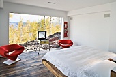 White bedroom with red designer armchairs in front of a large bank of windows with a view of the countryside