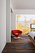 A red designer armchair in front of floor-to-ceiling windows in a bedroom with a view of the countryside