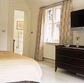 A white bedroom with a dark wooden chest of drawers and a view into an en suite bathroom