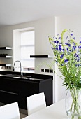 White dining area with blue summer flowers in front of a black modern kitchen with small floating shelves