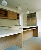 Kitchen unit in wood and light stone in front of simple, wooden wall-mounted shelving and a large, stainless-steel refrigerator