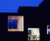 Zeitgenössisches Wohnhaus in Abendstimmung mit beleuchteten Fenstern und Blick auf Frau am Tisch