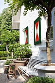 weiße Gartenbank und Tontöpfe mit Grünpflanzen vor Wohnhaus mit weisser Backsteinfassade und rotgrün bemalten Fensterläden