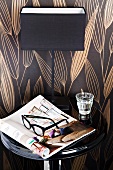 Still-life arrangement against chocolate-brown patterned wallpaper - newspaper, glasses, glass of water and elegant bedside lamp on black lacquered table