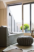 Simple, Scandinavian interior with pouffe in chunky knitted cover in front of light grey recamier and view of roof terrace with bright green box balls in background
