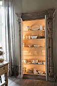 Crockery in illuminated, glass-fronted cabinet with open doors against grey-painted wall