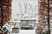 Schneebedeckter Arbeitstisch und Pflanzen vor weißer Hauswand
