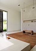 Ausgefallener Baderaum mit Zugang ins Grüne - künstlerisch gearbeiteter Holzhocker auf Holzrost vor schmaler Wandnische, im Vordergrund eine moderne Badewanne