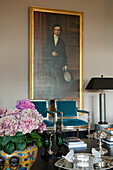 Painting of a man above armchairs with upholstery in petrol, table lamp and hydrangeas