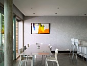 Colourful painting in grey room - long metal dining table and white plastic chairs next to kitchen counter with bar stools