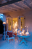 Festive atmosphere on veranda with lanterns and candlesticks on table in front of open living room door and view of illuminated Christmas tree
