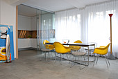 A dining table with lemon-yellow designer chairs in front of floor-to-ceiling windows and a white kitchenette