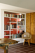 Open wall of books with red background, designer chair, rustic wooden table and tiled floor