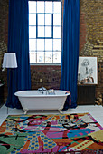 Loft bathroom with colourful rug and free-standing vintage bathtub in front of blue curtains