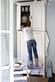 Woman standing on English garden chair putting away crockery in white, country house cupboard