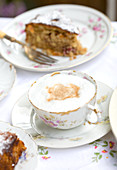 A cappuccino in an antique floral teacup surrounded by blurred plates of cake