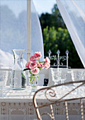 Roses in stemmed glass next to carafe of water and retro wine glasses on terrace table in front of translucent curtain