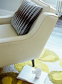 Cushion on armchair upholstered in white leather next to open book and mug on rug