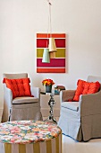 View over pouffe of orange cushions on armchairs with grey loose covers in front of wall with modern artwork and fifties pendant ceiling lamps