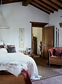Bett mit weisser Tagesdecke und Korbsessel im schlichten Schlafraum mit offen stehender Zimmertür