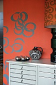 Chest of drawers painted light grey against red wall with retro-style circular pattern
